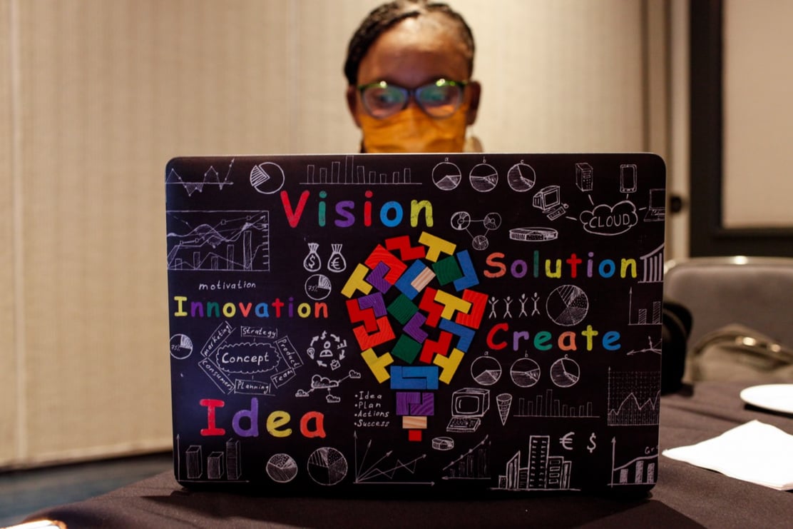 2022 DLF Forum attendee seated at their laptop. There is an illustration superimposed on the laptop cover with a colorful lightbulb and the words: vision, solution, innovation, create, idea